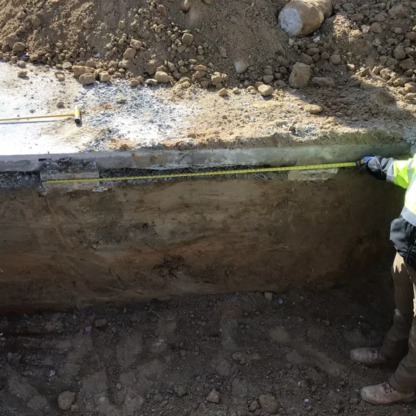 Construction worker in safety gear measuring trench depth beside concrete foundation and dirt mound.