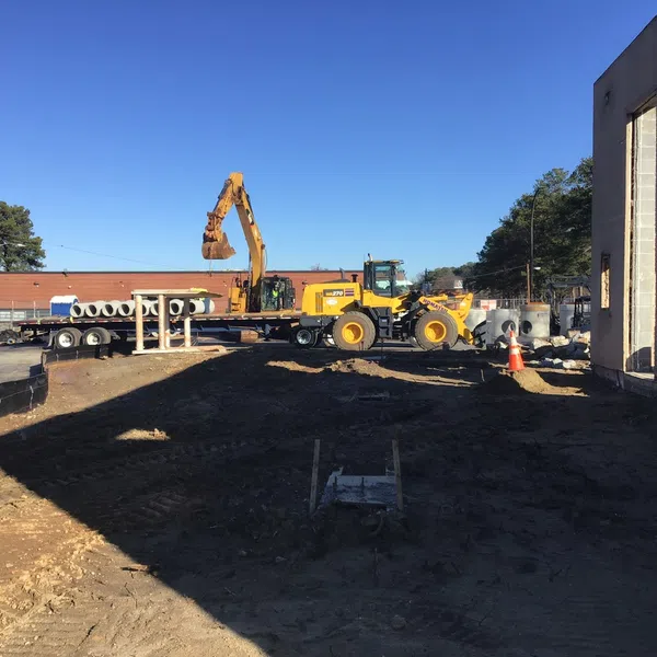 Construction site with excavator and loader working near brick building under clear blue sky.