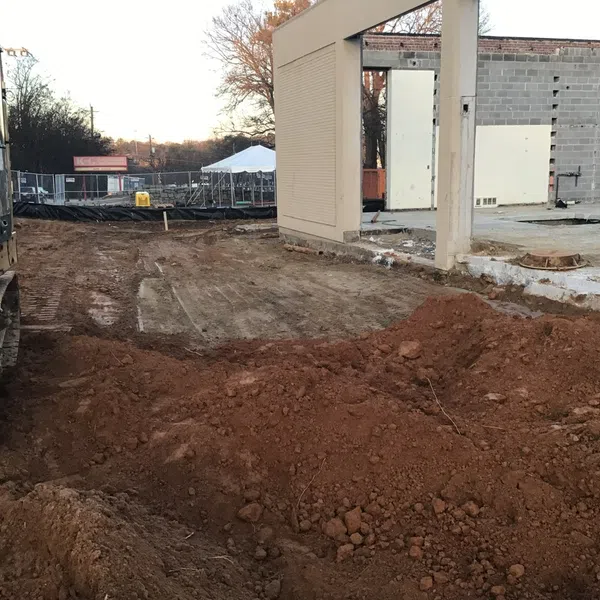 Construction site with excavator and dirt piles near a partially demolished building in the late afternoon.