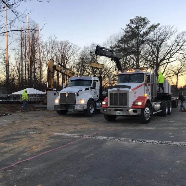 Two flatbed trucks with cranes operating at a construction site during sunset with workers and trees in background