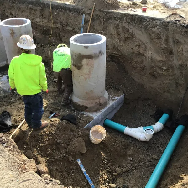 Construction workers install large concrete pipes and blue drainage pipes in a deep trench at a worksite.