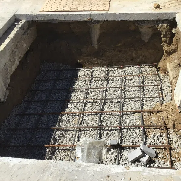 Excavated square foundation pit with gravel and steel rebar grid for concrete pouring.
