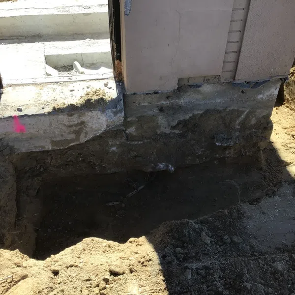 Excavation trench beside house foundation exposing soil and concrete for construction or repair work.