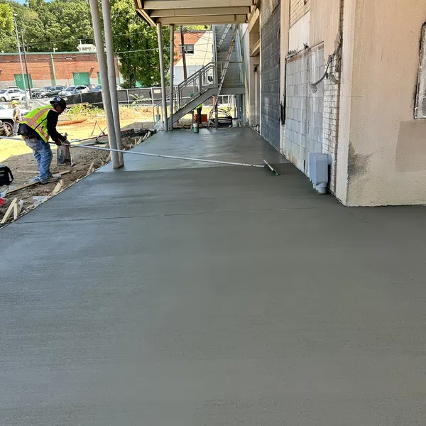 Construction worker smoothing fresh concrete sidewalk next to building with stairs under a metal awning