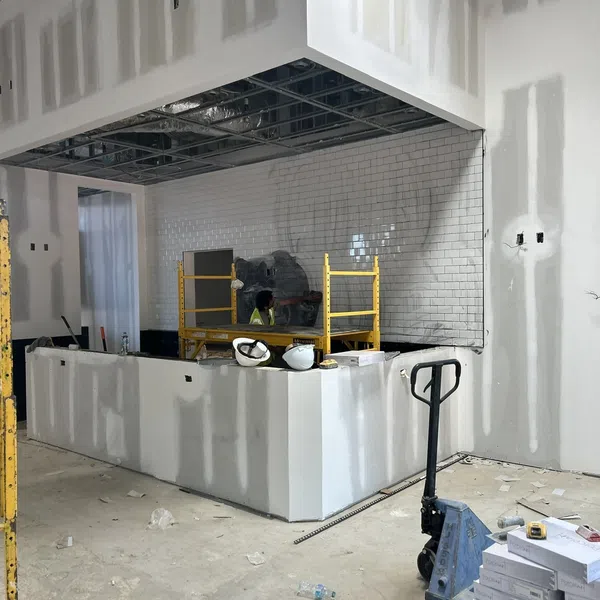 Interior under construction with drywall, white tiled wall, scaffolding, construction tools, and boxes on concrete floor.