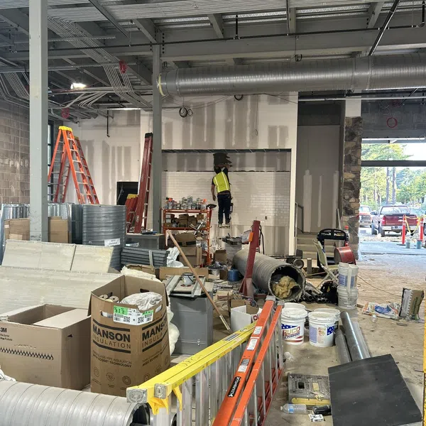 Construction site indoor with ladders, ducts, insulation materials, and a worker installing drywall or tiles.
