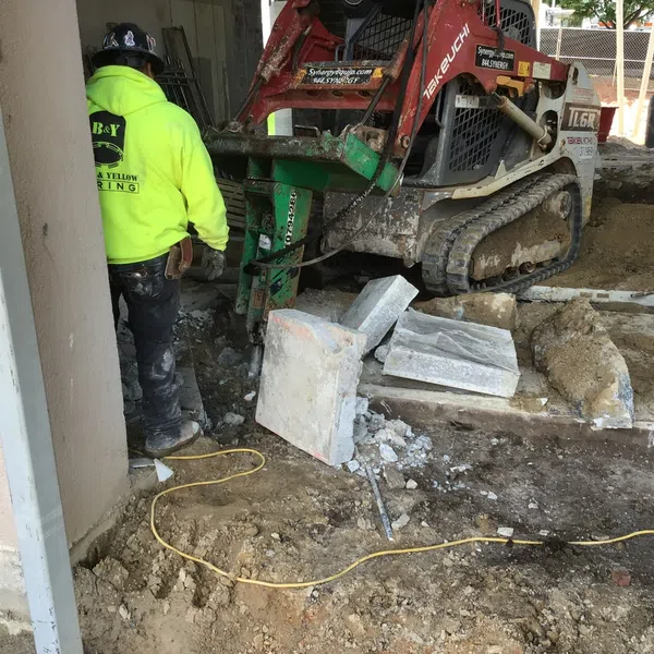 Construction worker operates compact excavator breaking concrete slabs on a commercial site under renovation.