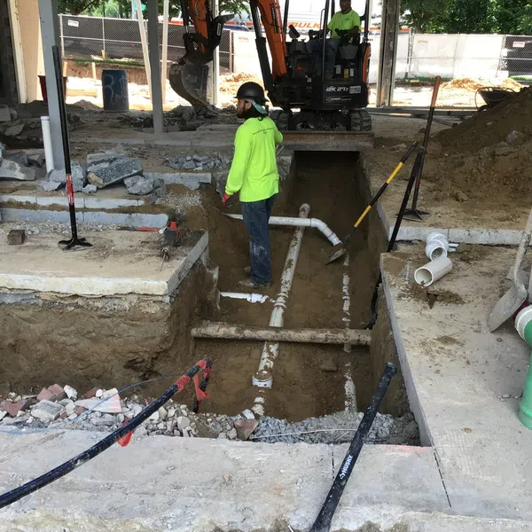 Construction site with workers installing underground PVC plumbing pipes and trench excavation under a building foundation.