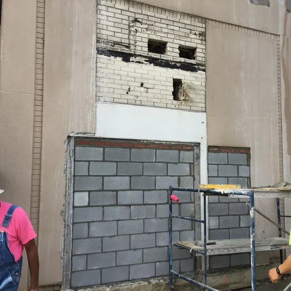 Workers in safety gear constructing a brick wall on a building exterior with scaffolding and cement blocks.