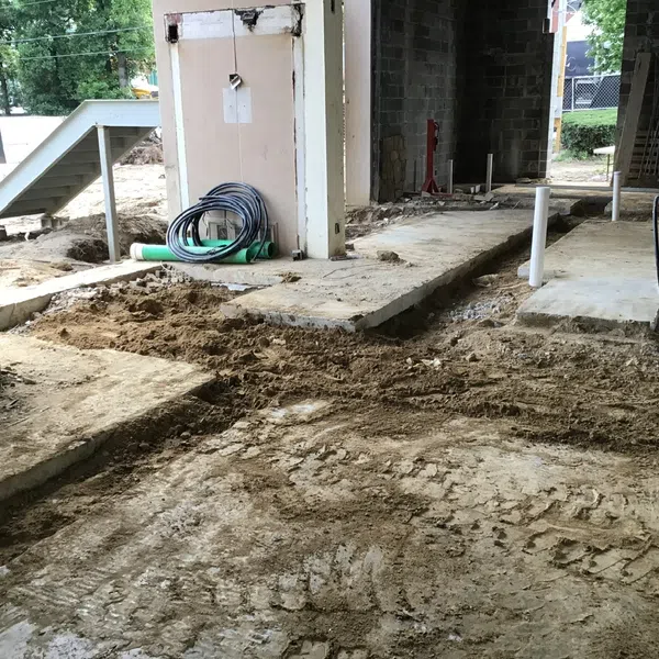 Construction site with exposed dirt floor, concrete slabs, pipes, and coiled cables inside an unfinished building.