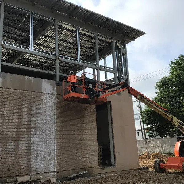 Construction worker on orange aerial lift working on steel frame of building with brick wall and trees in background