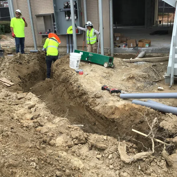 Construction workers in safety gear working around a deep trench with pipes and tools near a building site.
