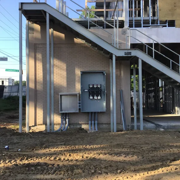 Construction site with metal staircase, electrical panels mounted on brick wall, and dirt ground in daylight