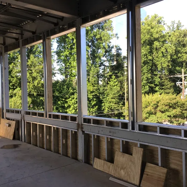 Interior view of a building under construction with metal framing and plywood panels, surrounded by green trees.