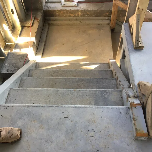 View down unfinished concrete stairs in a construction site with wooden framing and a construction boot visible.