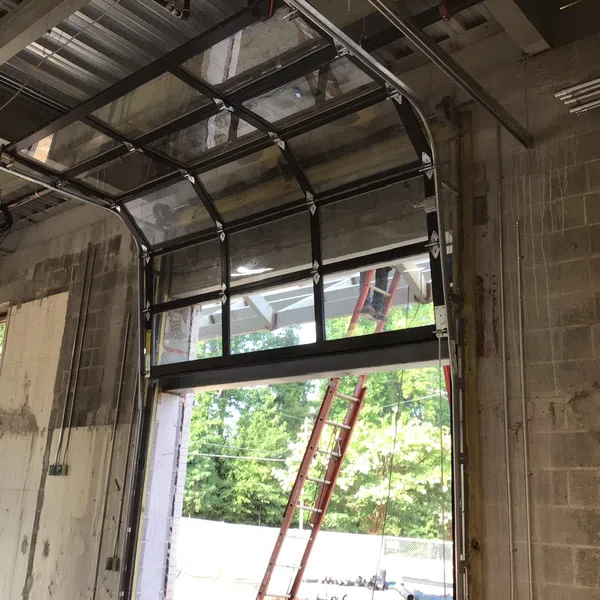 Industrial garage door partially open with metal frame and glass panels, showing an outdoor ladder and greenery.
