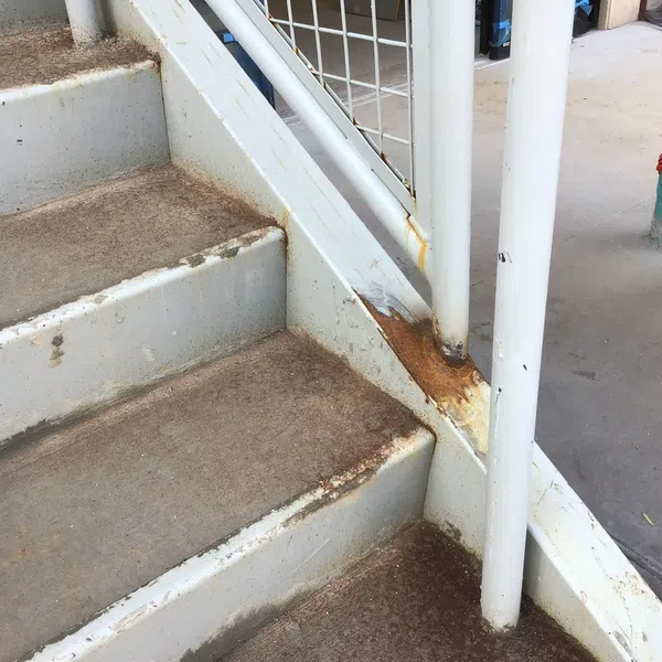 Close-up of rusty, worn metal stairs with white railings and peeling paint on concrete flooring.