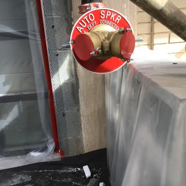 Red automatic sprinkler connection mounted on exterior wall near plastic-covered equipment at construction site