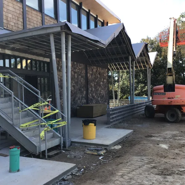 Construction site showing a building entrance with covered walkway, stairs, and orange hydraulic lift.