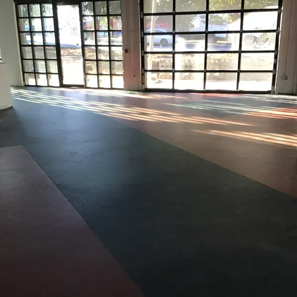 Empty industrial space with large glass garage doors and sunlight casting shadows on multicolored floor.