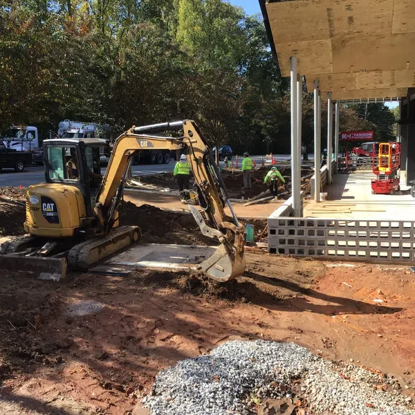 Small excavator digging dirt at a construction site near a commercial building with workers in safety vests.