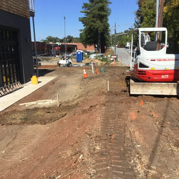 Construction site with a Takeuchi excavator, dirt ground, traffic cones, and building under clear blue sky.