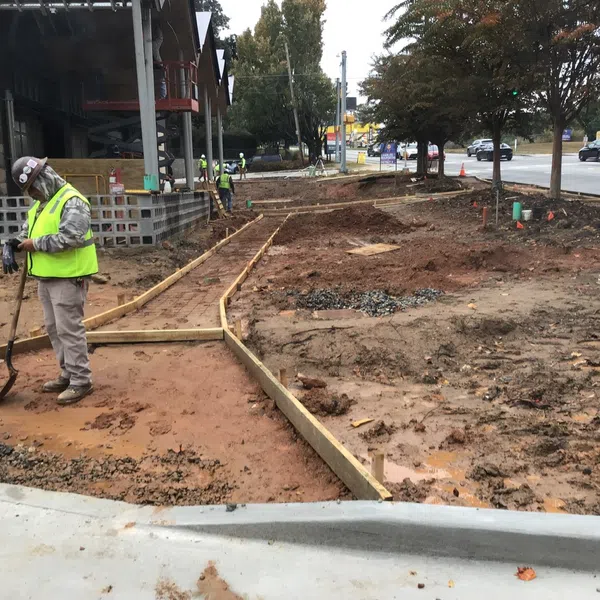 Construction workers in safety vests preparing ground and wooden framework for new sidewalk along a busy street.