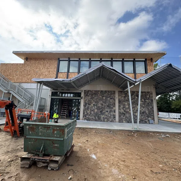 Modern building under construction with stone wall, metal framework awnings, and construction equipment on site.