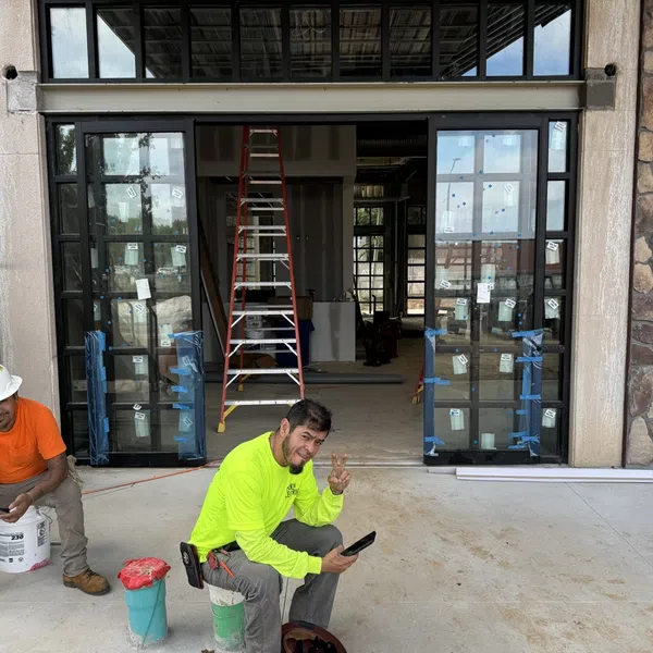 Two construction workers resting outside a building entrance with glass doors and ladder inside, one making a peace sign.