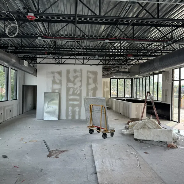 Interior of a commercial space under construction with exposed ceiling, drywall, large windows, and construction materials.