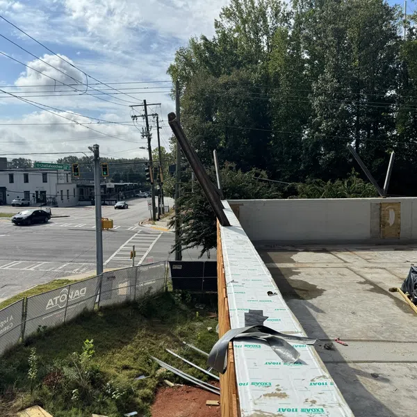 Partially constructed building roof near busy intersection with trees and cars under blue sky with clouds.
