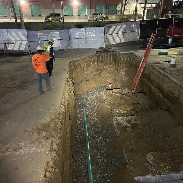 Two construction workers inspect a deep trench with pipes and a red ladder at a dimly lit night construction site.