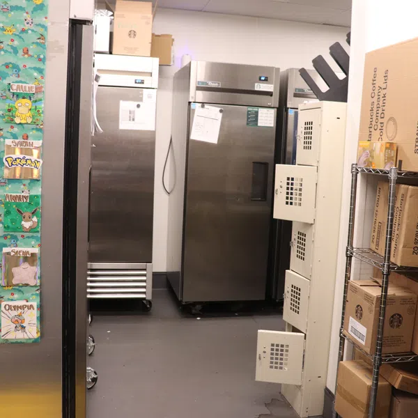 Storage room with metal refrigerators, cardboard boxes on a metal shelf, and a colorful bulletin board with name tags.