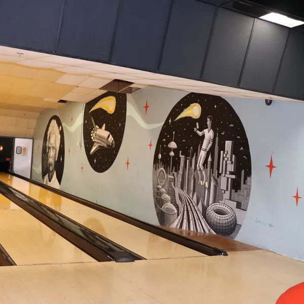 Bowling alley lanes next to a wall with futuristic and space-themed murals and a red bowling ball in the foreground.
