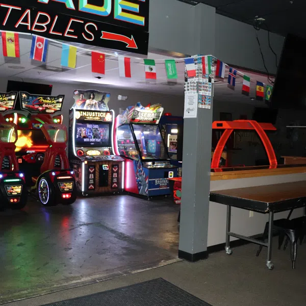Arcade gaming area with illuminated racing and fighting game machines, decorated with international flags overhead.