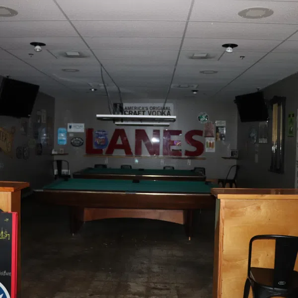 Dimly lit pool hall with two tables, bar stools, wall TVs, and signage including 'LANES' and pool table rules.