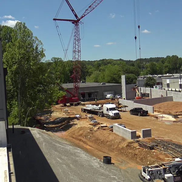 Construction site with cranes, trucks, and building materials surrounded by trees and industrial buildings under blue sky.