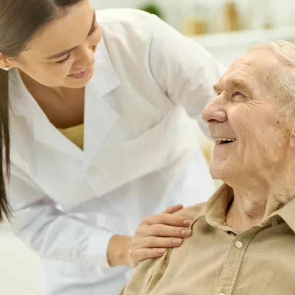 Elderly man smiling with a young female caregiver in a supportive, warm interaction indoors.