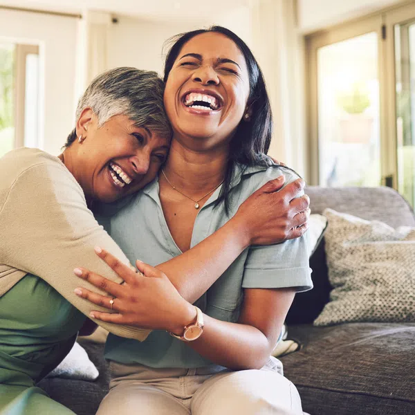 Two women sharing a joyful hug and laughter while sitting on a couch in a bright living room.