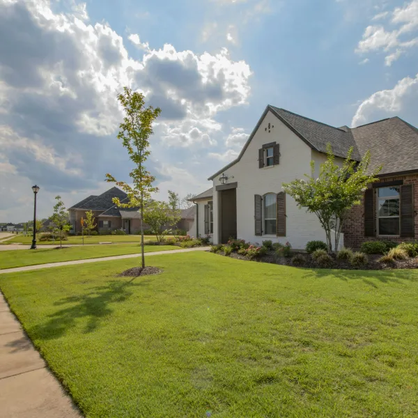 Suburban house with brick and white exterior, manicured lawn, young trees, and a cloudy blue sky.
