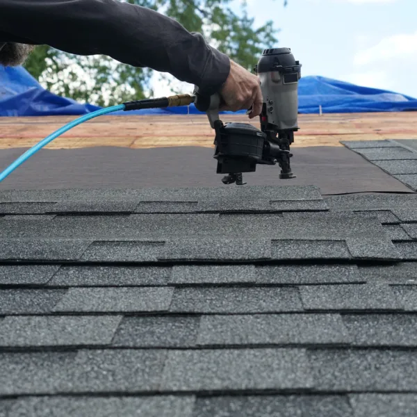 Worker using nail gun to install asphalt shingles on a roof under a blue sky.