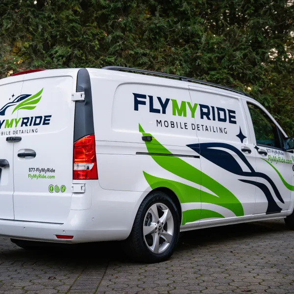 White Fly My Ride mobile detailing van with green and black graphics parked on a paved driveway surrounded by trees.