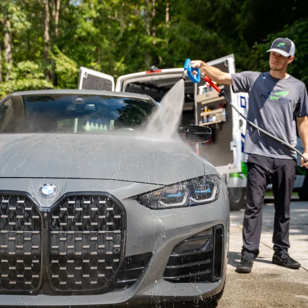 Man washing a gray BMW car outdoors with a high-pressure spray from a van in the background.