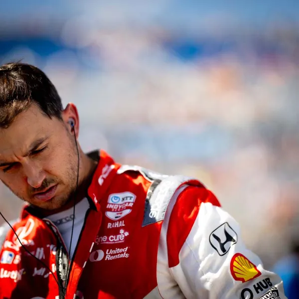 Race car driver in red and white suit adjusting earphones before race with blurred background.