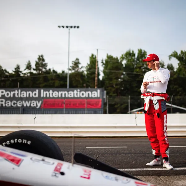 Race car driver in red and white suit stands near his car at Portland International Raceway.