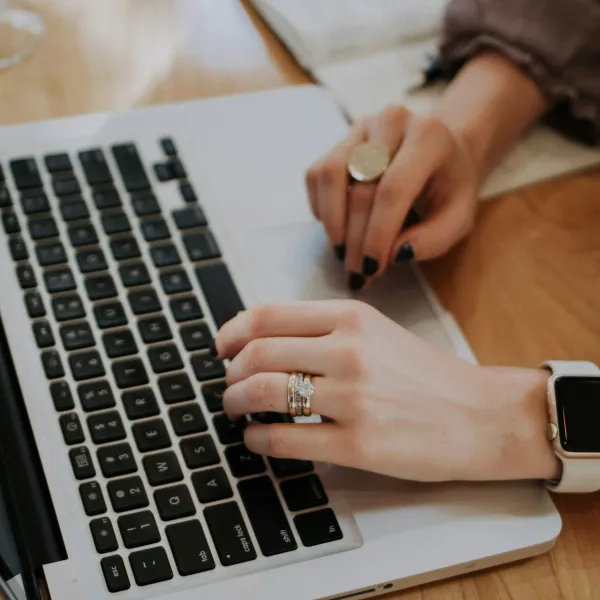 Person typing on a laptop keyboard with rings and a smartwatch on wooden table background
