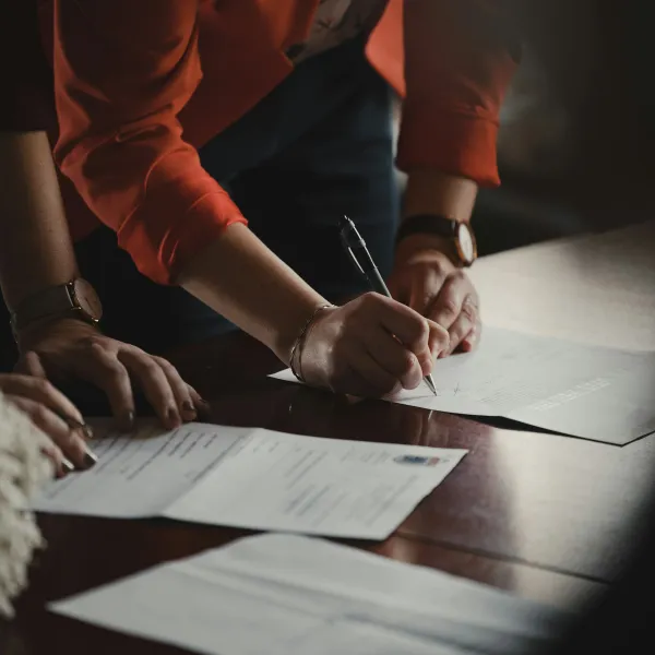 Person signing documents on a wooden table with another individual overseeing the process.