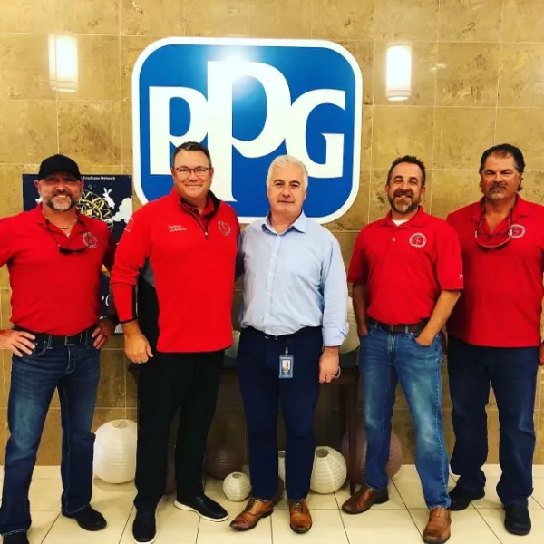 Five men standing in front of a large PPG logo on a beige tiled wall, four in red shirts and one in a blue shirt.