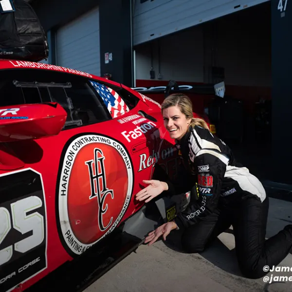 Smiling female race car driver kneeling next to red race car number 125 in garage area.
