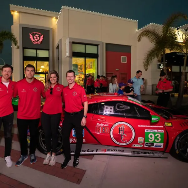 Four people in red shirts posing beside a red racing car at night outside a building with palm trees and people.